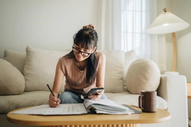 Asian woman planning budget and using calculator on smartphone. A woman sits at her living room with smartphone and financial reports doing her monthly budget. invoice stock pictures, royalty-free photos & images
