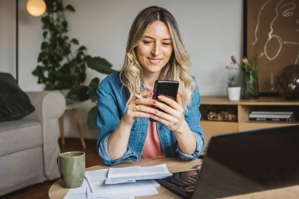 Working at home. Young woman going through paperwork in the living room at home. She is using smart phone while working on laptop. invoice stock pictures, royalty-free photos & images
